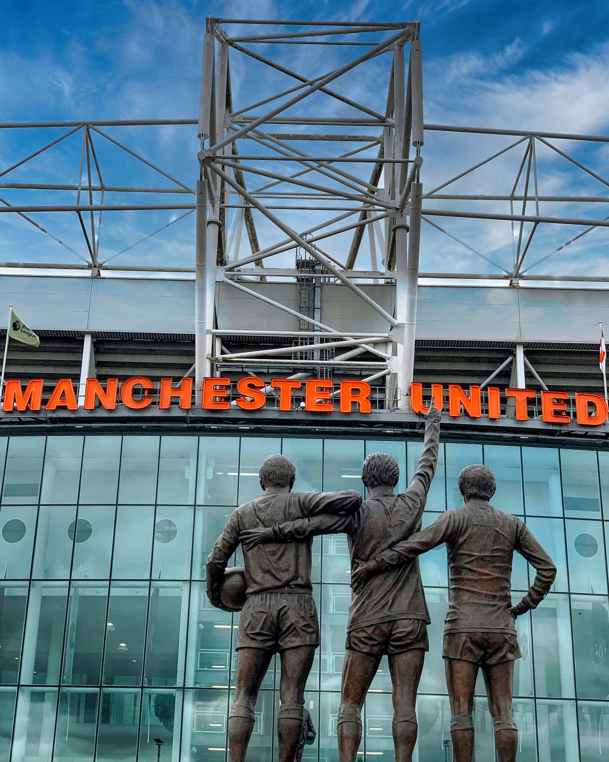 The iconic United Trinity statue stands beneath the vibrant orange "MANCHESTER UNITED" lettering at Old Trafford stadium, a must-see highlight of the stadium tour experience.